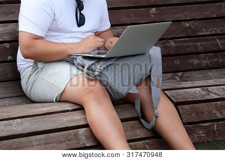 Fat Man In Shorts Sitting With A Laptop On A Bench In A Summer City. Male Hands On A Keyboard, Conce