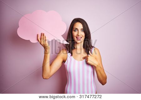 Beautiful woman wearing striped pink swimsuit and holding talking balloon over pink background with surprise face pointing finger to himself
