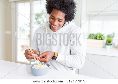 African American man eating asian noodles using chopsticks at home with a happy face standing and smiling with a confident smile showing teeth
