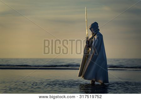 Portrait Of Assassin In White Costume With The Sword At The Sea. He Is Posing Near Water During Suns