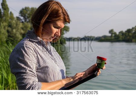 Woman On River Bank Entering Data On Tablet About Green Algae Taken In Water