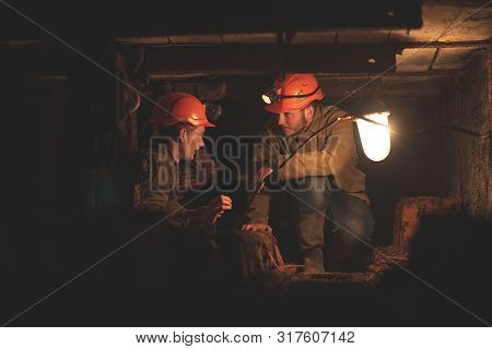 Two Young Guys In A Working Uniform And Protective Helmets, Sitting In A Low Tunnel. Workers Of The