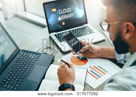 Bearded Man Freelancer Sits In Office At Table, Uses Smartphone, Working On Laptop With Inscription