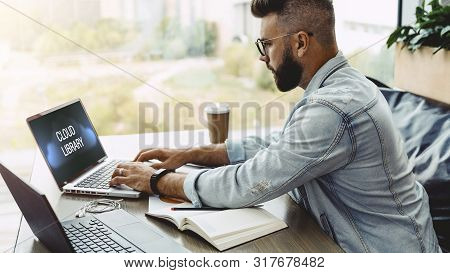 Bearded Man Hipster Sits In Cafe At Table And Works On Laptop With Inscription On Monitor- Cloud Lib