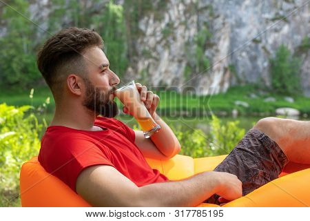 A Young Man With A Beard Lies On An Orange Inflatable Sofa Lamzak And Drinks Beer From A Glass On Th