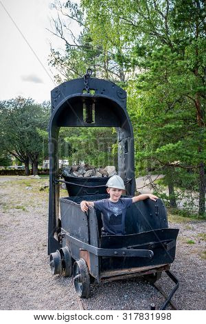Outdoor Portrait Of Young Child Sitting In An Old Black Iron Mining Carriage Wagon Wearing A White H