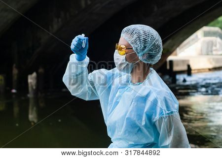 Female Ecologist Or Epidemiologist Takes Test-tube Water Analysis In A City River Under An Old Bridg