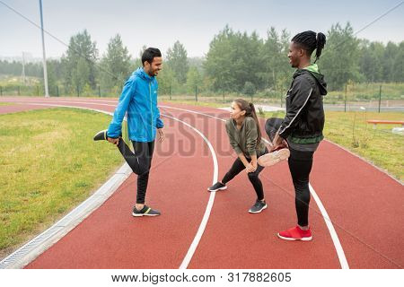 Group of young intercultural athletes doing wam-up exercises on racetracks