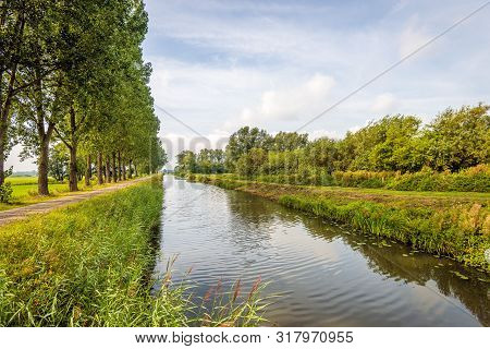 Long And Straight Canal In A Rural Landscape In The The Dutch Summer Season. Along The Canal Is A Ro