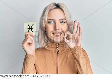 Young blonde girl holding paper with pisces zodiac sign doing ok sign with fingers, smiling friendly gesturing excellent symbol 
