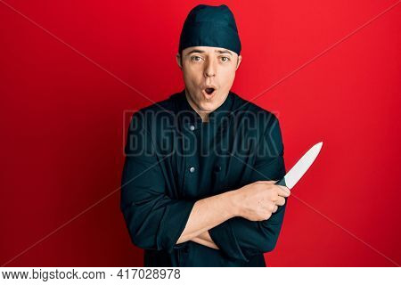 Handsome young man wearing professional cook uniform holding knife afraid and shocked with surprise and amazed expression, fear and excited face. 