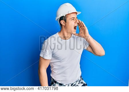 Handsome young man wearing builder uniform and hardhat shouting and screaming loud to side with hand on mouth. communication concept. 