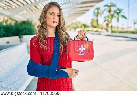 Young blonde girl with serious expression injuried with arm sling and holding first aid kit at the park.