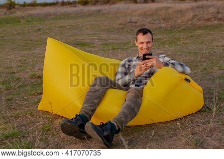 Young Man Sitting On Inflatable Sofa Lamzac With Smartphone While Resting At Sunset On The Mountain
