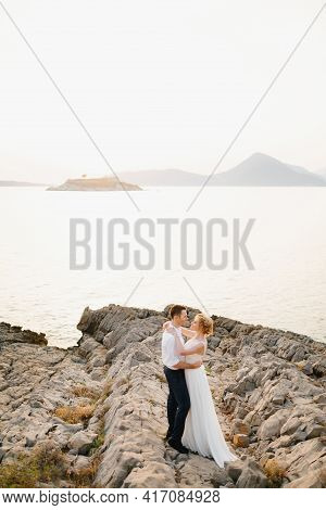 The Bride And Groom Hug On The Rocks By The Sea Against The Backdrop Of The Mountains And The Island