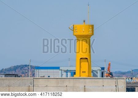 Yellow Lighthouse On End Of Concrete Pier With Buildings In Background.