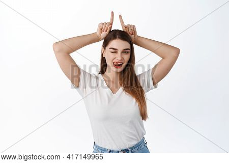 Sassy And Flirty Young Woman Wink, Shows Tongue And Bull Devil Horns Gesture, Fingers On Head, Being