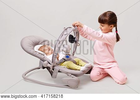 Baby Girl Lying Down In Cradle With Her Sister, Little Girl With Pigtails Sitting On Floor Near Her 