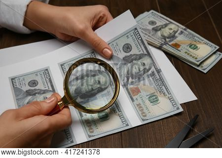 Counterfeiter Examining Sheet Of Paper With Dollar Banknotes At Wooden Table, Closeup. Fake Money Co