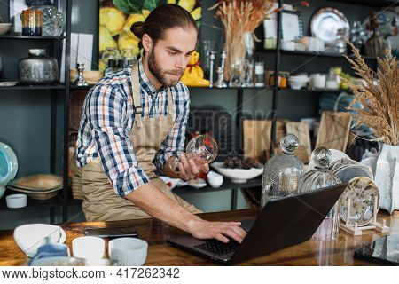 Bearded Man In Uniform Sitting At Decor Store And Working On Modern Laptop. Competent Seller Recheck