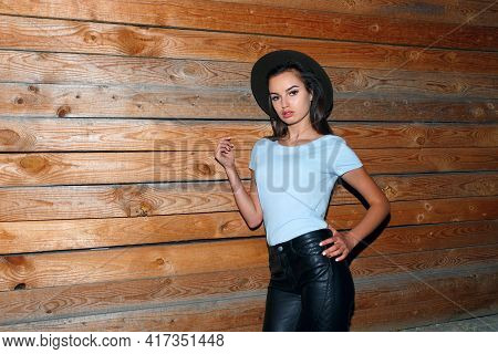 Portrait Of A Female Model In Hat, Her Hair Straight Dressed In Leather Pants And Blue T Shirt Near 