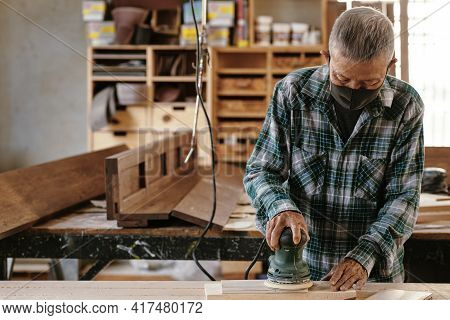 Elderly Experienced Carpenter In Protective Mask Polising Wooden Plank With Electric Sander