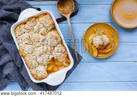 Serving Of Freshly Baked Drop Biscuit Peach Cobbler With Apron And Wooden Spoon Shot From Top View.