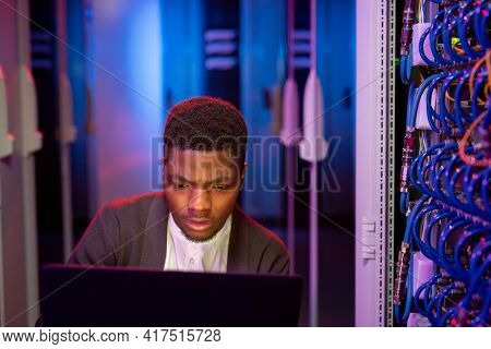 Busy young black IT engineer using modern laptop in server room while administrating computer network