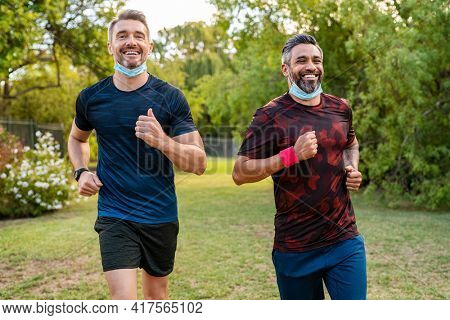 Happy mature man with indian friend running at park with surgical face mask during covid-19 pandemic. Active mid adult man jogging outdoor with lowered face mask against coronavirus.
