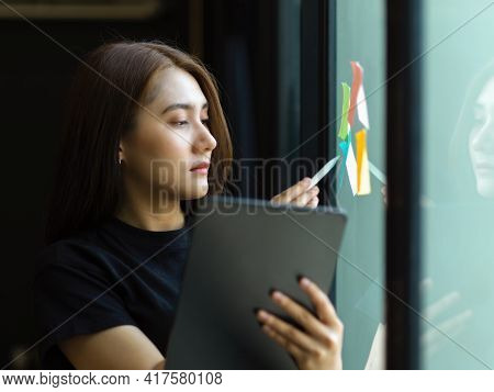 Female Office Worker Working With Sticky Note With Idea On Glass Wall And Digital Tablet