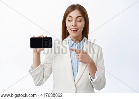 Smiling Business Woman In Professional Suit, Points At Empty Smartphone, Holds Screen Horizontal, In