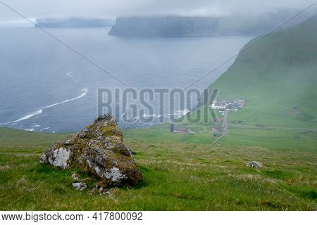 Faroese Village Of Trollanes Viewed From A Hill Nearby. Green Hills, Cloudy, Misty Weather In The Wi