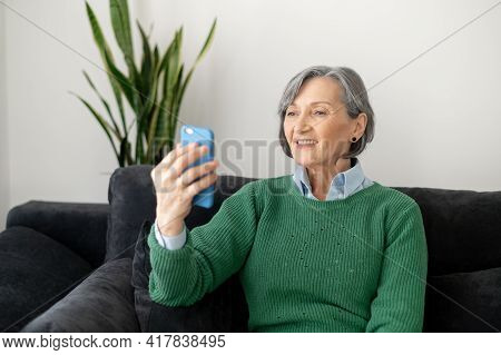 Excited Senior Mature Lady Wearing Green Sweater Sitting On The Couch In The Living Room, Holding A 