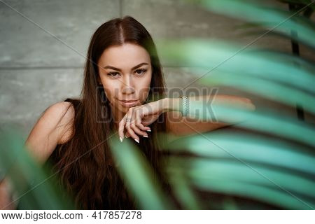 Beautiful Young Woman Closeup Portrait Through Green Leaves Of A Flower On A Gray Wall Background