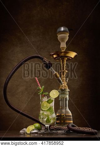 Hookah (shisha) and glass of Mojito cocktail with lime and ice on dark brown wooden table on dark background. Weekend or holiday party