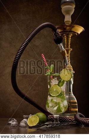 Hookah (shisha) and glass of Mojito cocktail with lime and ice on dark brown wooden table on dark background. Weekend or holiday party