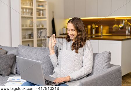 Happy Young Woman Sits On The Comfortable Sofa With A Laptop And Has Video Call, Waving Hello Into W