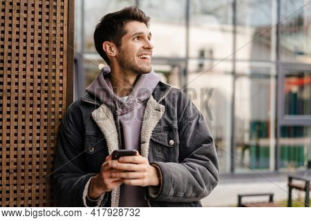 Smiling young white man in casual cothes using mobile phone outdoors in the city, leaning on a wall