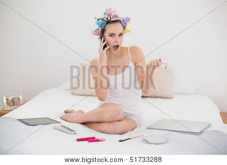 Astonished natural brown haired woman in hair curlers calling with her mobile phone in bright bedroom