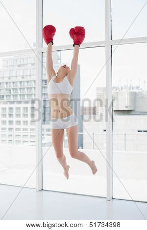 Cheerful fit brown haired model in sportswear jumping and wearing red boxing gloves in bright fitness studio