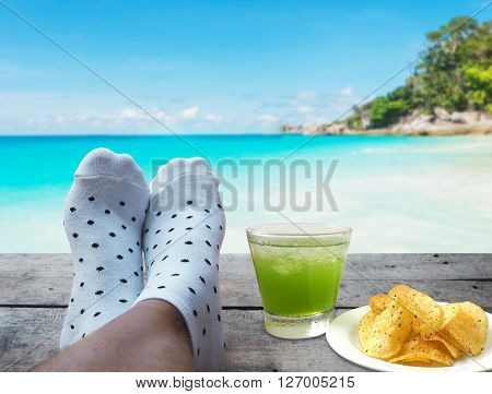 feet on wooden floor with glass of apple juice and potato chips over beach background Relaxtion on vacation concept
