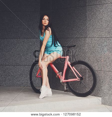 Sexy girl on bicycle. Young slim sexy sportive woman in blue shorts and white snickers long-haired, sensual posing on pink fix bicycle at grey background in urban city enviroment. Soft toned.