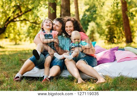 Young happy family with kids having picnic with colored pillows outdoors. Parents with two children relax in a sunny summer garden. Mother father little girl and baby boy playing in park.