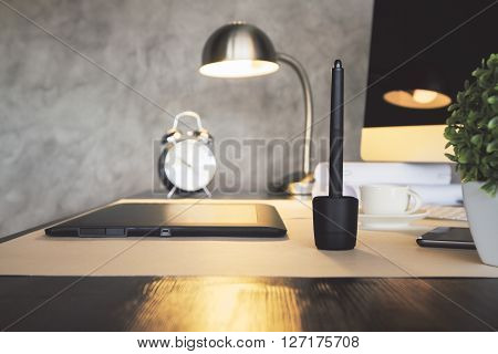 Sideview of desktop with graphic tablet plant alarm clock illuminated lamp and computer. Concrete wall background