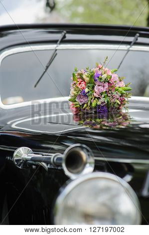Elegant bridal bouquet of different flowers on the hood of a black retro car. Lacquered surface reflection. Windshield in the background. Headlight and retro car horn in front. Focus on a bouquet.