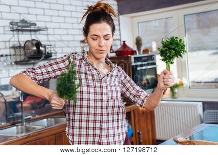 Happy young woman with eyes closed holding dill and parsley and dacing on the kitchen