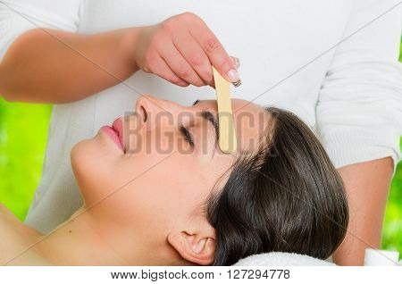 Closeup womans face receiving facial hair waxing treatment, hand using wooden stick to apply wax, beauty and fashion concept.
