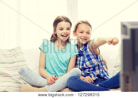 people, children, television, friends and friendship concept - two happy little girls watching tv and pointing finger at home