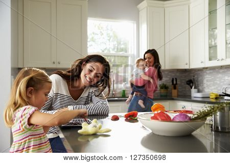 Girl uses tablet in kitchen with mum, other mum holding baby