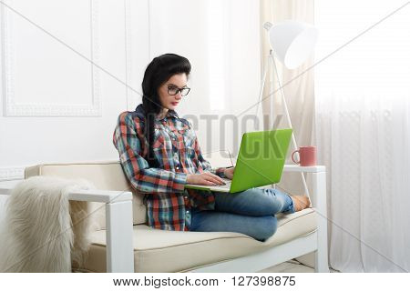Young girl in protective computer glasses on a white sofa with laptop. Hipster freelancer woman works at home. Home office, freelance. Brunette girl with laptop in modern interior at white couch.